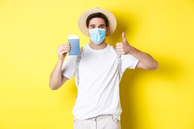 Concept of covid-19, tourism and pandemic. Happy male tourist in medical mask showing passport, going on vacation during coronavirus, make thumb up sign, yellow background.