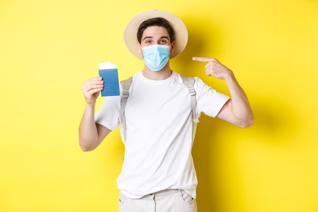 covid-19, 관광 및 전염병의 개념. 코로나바이러스로부터 보호하기 위해 의료 마스크를 쓰고 여행하는 여권을 보여주는 남자 관광객, 노란색 배경