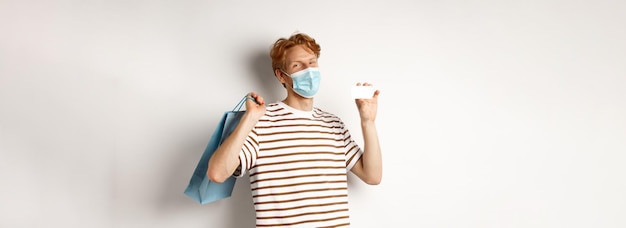 Free photo concept of covid and lifestyle happy young shopper in face mask holding shopping bag and showing pla