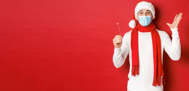 パンデミック中のcovid19クリスマスと休日の概念医療用マスクとサンタ帽子をかぶった陽気なハンサムな男は、線香花火で新年を祝い、赤い背景を興奮させました