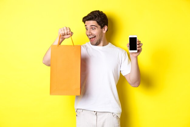 Concept of discounts, online banking and cashback. Happy guy buy something in store and looking at shopping bag, showing mobile phone screen, yellow background.