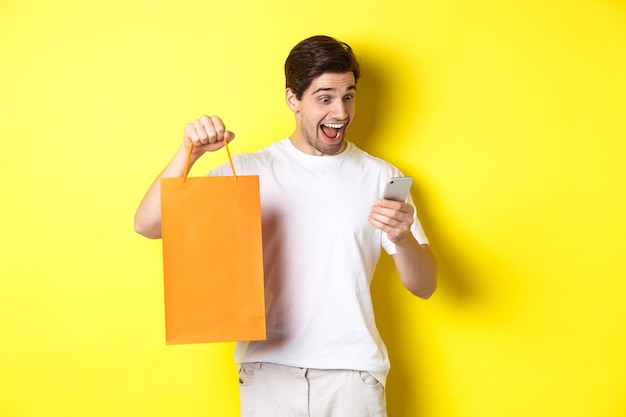 Концепция скидок, интернет-банкинга и кэшбэка. Удивленный человек показывает хозяйственную сумку и смотрит счастливым на мобильный экран, стоя на желтом фоне.