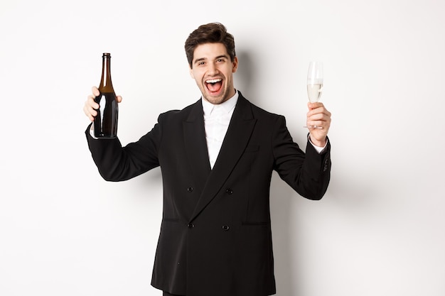 休日、パーティー、お祝いの概念。楽しんで、白い背景の上に立って、シャンパンのボトルとグラスを持って、トレンディなスーツを着たハンサムな男。