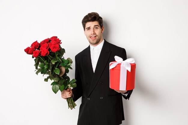 Concept of holidays, relationship and celebration. Handsome and confident man in black suit, going on a date, holding bouquet of roses and present, standing against white background