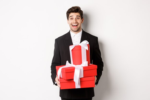 Concept of holidays, relationship and celebration. Handsome man in black suit bringing presents at new year party, holding gifts and smiling amused, standing against white background.