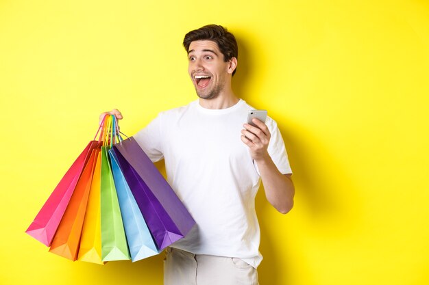 Concept of mobile banking and cashback. Happy man looking amazed, holding shopping bags and smartphone, yellow background.