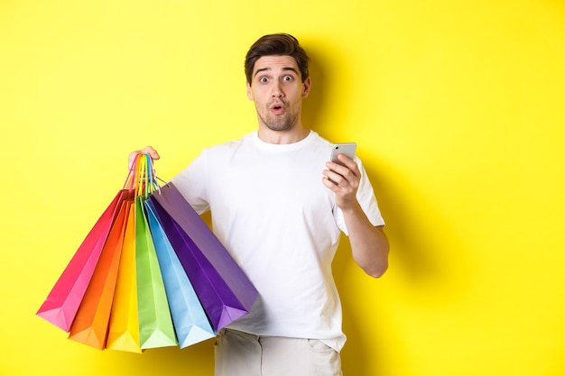 Concept of mobile banking and cashback. Surprised man holding shopping bags and smartphone, standing over yellow background