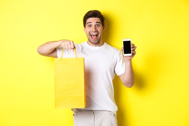 모바일 뱅킹 및 캐쉬백의 개념. 쇼핑백을 들고 스마트 폰 화면, 노란색 배경 보여주는 젊은 행복 한 남자.