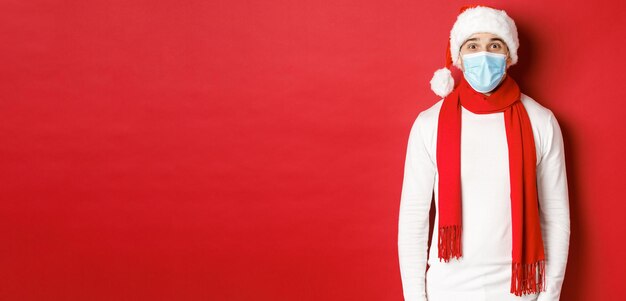 新年、コロナウイルス、休日の概念。クリスマスと社会的な距離を祝う陽気な男、医療マスク、サンタの帽子とスカーフを身に着けて、赤い背景の上に立っています。