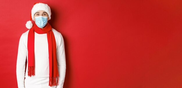 새해, 코로나바이러스 및 휴일의 개념. 크리스마스와 사회적 거리를 축하하는 쾌활한 남자, 의료용 마스크, 산타 모자, 스카프를 착용하고 빨간색 배경 위에 서 있습니다.