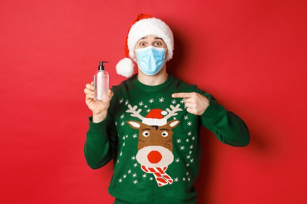 新年のコンセプト、コロナウイルスと社会的距離。サンタ帽子、クリスマスセーター、医療マスク、手指消毒剤、赤い背景の上に立っている陽気な男の肖像画。