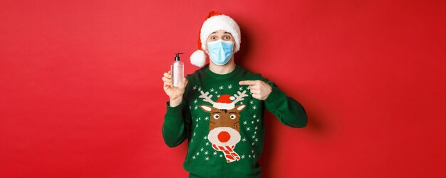 새해, 코로나바이러스, 사회적 거리의 개념. 산타 모자, 크리스마스 스웨터, 의료 마스크를 쓴 쾌활한 남자의 초상화, 손 소독제를 손가락으로 가리키며 빨간색 배경 위에 서 있는