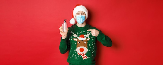 Concept of new year, coronavirus and social distancing. Portrait of cheerful man in santa hat, christmas sweater and medical mask, pointing finger at hand sanitizer, standing over red background