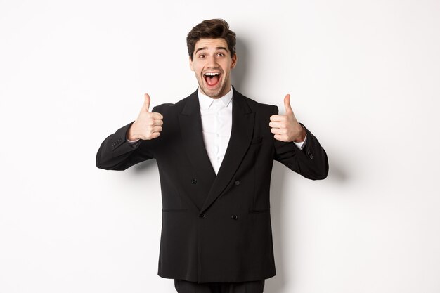 Concept of new year party, celebration and lifestyle. Portrait of amazed and pleased handsome man in black suit, showing thumbs-up, like product, approve something good, white background