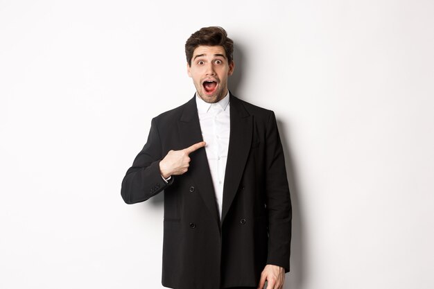 Concept of new year party, celebration and lifestyle. Portrait of surprised handsome guy in black suit, pointing at himself and looking amazed, being chosen, standing over white background