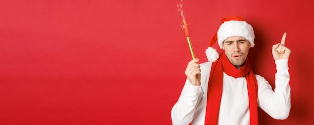 크리스마스, 겨울 방학 및 축하의 개념입니다. 새해 파티를 즐기고, 스파클러와 함께 춤을 추고, 산타 모자와 스카프를 쓰고, 빨간 배경 위에 서 있는 매력적인 남자.