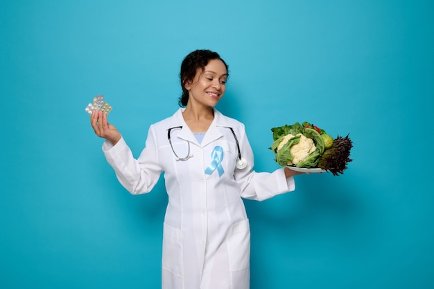 적절한 영양과 건강 관리의 개념입니다. 파란색 당뇨병 인식 리본이 달린 의료 가운을 입은 자신감 있는 여성 의사는 건강한 채식주의 음식 한 접시와 약이 담긴 물집을 비늘로 들고 있습니다.