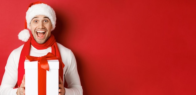 Бесплатное фото Концепция зимних праздников, рождества и образа жизни крупный план возбужденного красивого мужчины в санта-джате и шарфе, выглядящего изумленным и получающего новогодний подарок, стоя на красном фоне