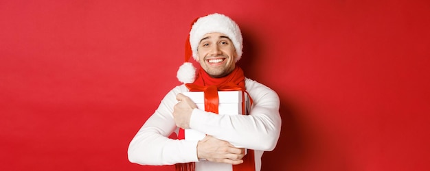 冬の休日、クリスマス、ライフスタイルのコンセプト。サンタの帽子とスカーフに身を包んだ素敵な男の画像。新年のプレゼントを抱きしめ、赤い背景の上に立って、お世辞を言って笑っています。