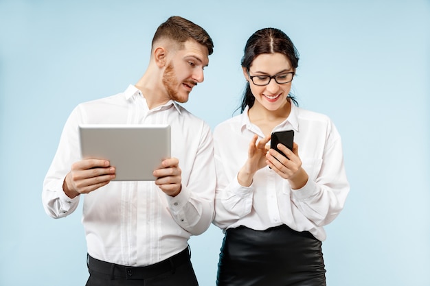 Concept of partnership in business. Young happy smiling man and woman standing with phone and tablet against blue background at studio