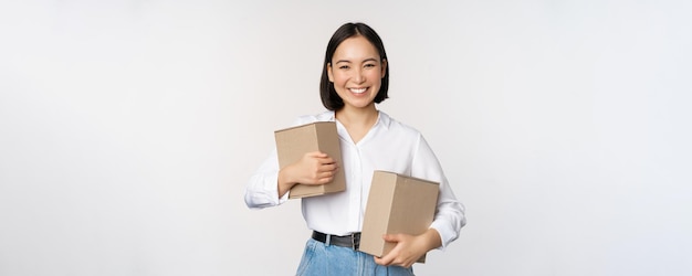 쇼핑 및 배달의 개념 상자를 들고 포즈를 취하고 흰색 배경 위에 서서 웃고 있는 젊은 행복한 아시아 여성
