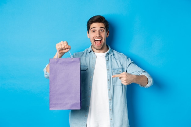 ショッピング、休日、ライフスタイルの概念。興奮した男が紙袋に指を向けて驚いたように見え、店を推薦し、割引を発表し、青い背景