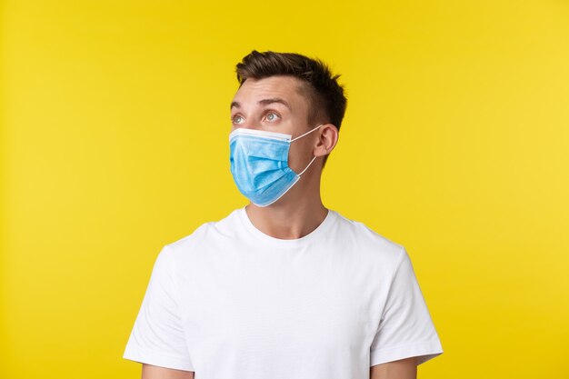 Concept of social distancing, covid-19 and people emotions. Close-up portrait of surprised and shocked blond guy in medical mask, raising eyebrows and staring speechless, yellow background.