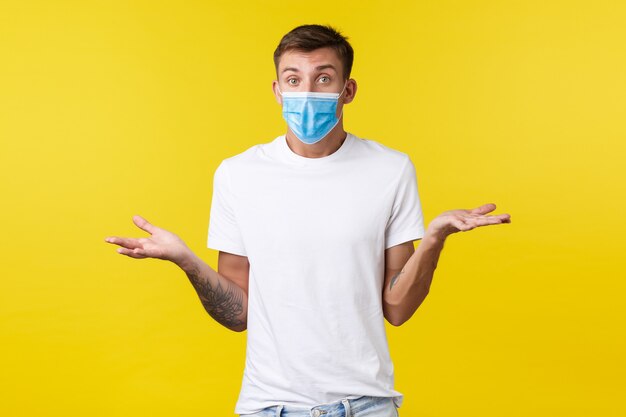 Concept of social distancing, covid-19 and people emotions. Confused and clueless handsome man in medical mask and basic t-shirt, shrugging unaware, have no idea, puzzled to answer, yellow background