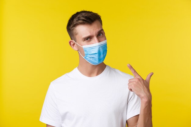 Concept of social distancing, covid-19 and people emotions. Handsome determined young man pointing finger at medical mask on face as recommend wearing eat outside, yellow background.