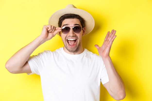 Concept of tourism and holidays. Close-up of surprised man shouting for joy, enjoying vacation, wearing sunglasses with summer hat, yellow background.