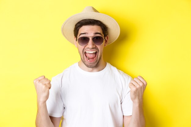 Concept of tourism and holidays. Lucky man tourist winning tickets, making fist pump and saying yes, wearing vacation clothes, standing over yellow background.