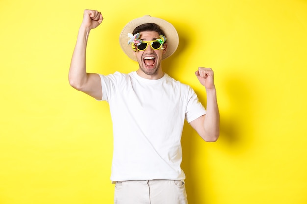 Понятие о туризме и образе жизни. Счастливый человек, выигравший поездку на курорт, кричит «да» и поднимает руки, торжествует, носит солнцезащитные очки и летнюю шляпу, желтый фон.