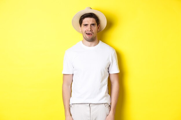 Concept of tourism and summer. Confused guy traveller in straw hat, looking puzzled, cant understand something, standing over yellow background