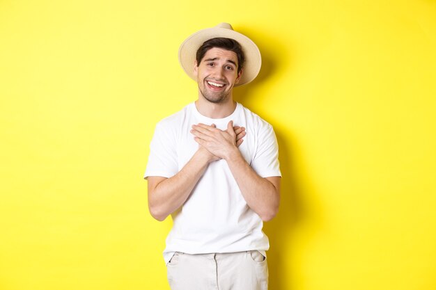 Concept of tourism and summer. Grateful guy in straw hat holding hands on heart, saying thank you and smiling with gratitude, standing against yellow background