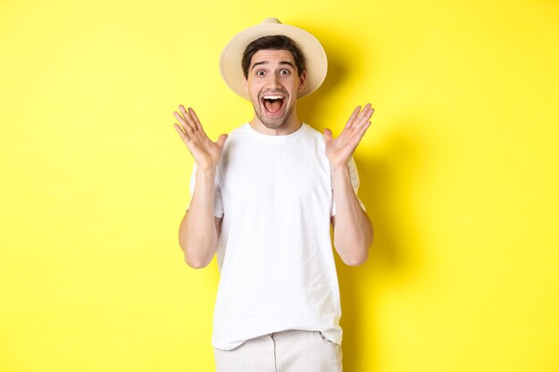 Concept of tourism and summer. Happy young man in straw hat looking amazed, reacting to surprise, standing over yellow background