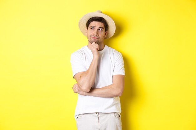 Concept of tourism and summer. Thoughtful man tourist pondering, looking at upper left corner and thinking, standing in straw hat and white t-shirt against yellow background.