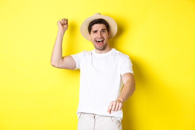 관광 및 여름의 개념입니다. 로데오 제스처를 보여주는 젊은 남자 여행자, 밀짚 모자와 흰색 옷을 입고 노란색 배경 위에 서