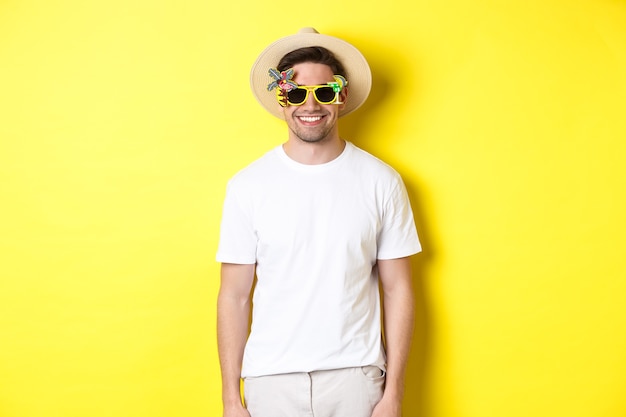 Concept of tourism and vacation. Relaxed smiling man enjoying supper trip, wearing sunglasses and straw hat, yellow background.