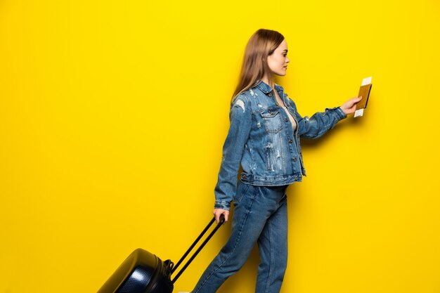 旅行のコンセプトです。黄色の壁でスーツケースとパスポートを持つ幸せな女少女