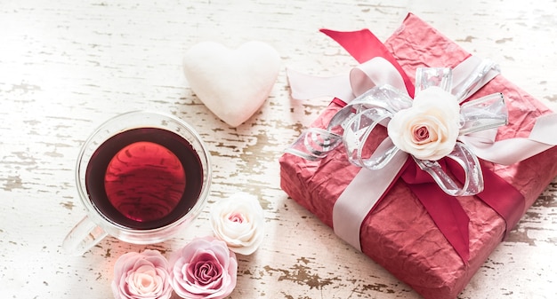 Free photo the concept of valentine's day and mother's day, a red gift box with a bow with roses and a cup of tea on a light wooden background