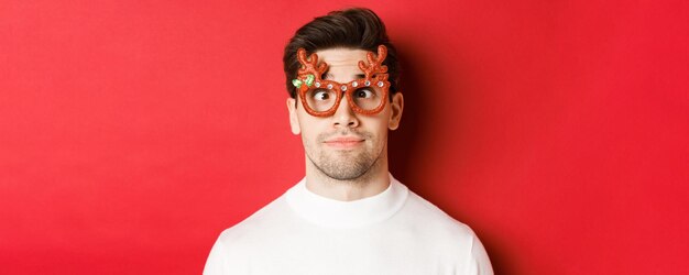 Concept of winter holidays, christmas and celebration. Close-up of funny brunette in party glasses, squinting and making faces, standing over red background.