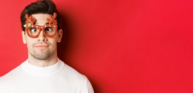 Concept of winter holidays christmas and celebration Closeup of funny brunette in party glasses squinting and making faces standing over red background