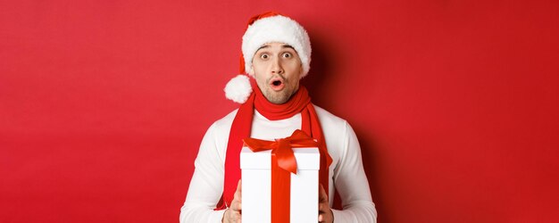 Концепция зимних праздников, Рождества и образа жизни. Крупный план удивленного красивого парня в новогодней шапке и шарфе, выглядящего изумленным и держащего новогодний подарок, стоящего на красном фоне.