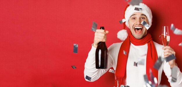 Концепция зимних праздников, рождества и образа жизни Крупный план счастливого человека, празднующего новый год, держащего бутылку шампанского и бокал, стоящий на красном фоне с конфетти