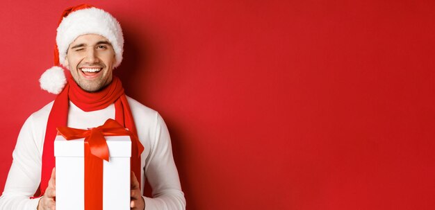 겨울 휴가, 크리스마스 및 라이프 스타일의 개념입니다. 산타 모자와 스카프를 쓴 건방진 잘생긴 남자, 선물을 들고 웃고, 카메라에 윙크하고, 빨간색 배경 위에 서 있는