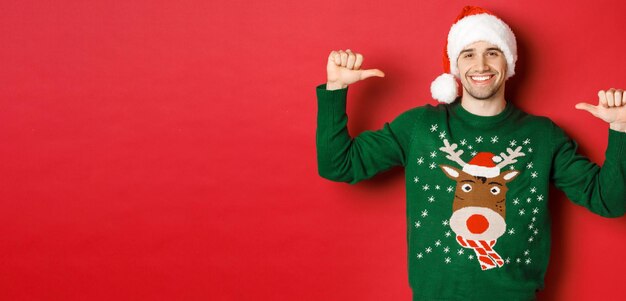 Concept of winter holidays christmas and lifestyle Happy attractive guy in santa hat and sweater pointing at himself with pleased face standing over red background