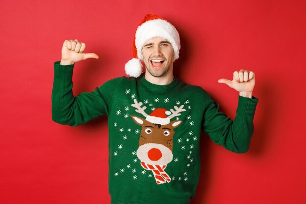 冬休み、クリスマス、ライフスタイルのコンセプト。サンタの帽子と緑のセーターを着た生意気なハンサムな男、自分を指してウインク、赤い背景の上に立っている
