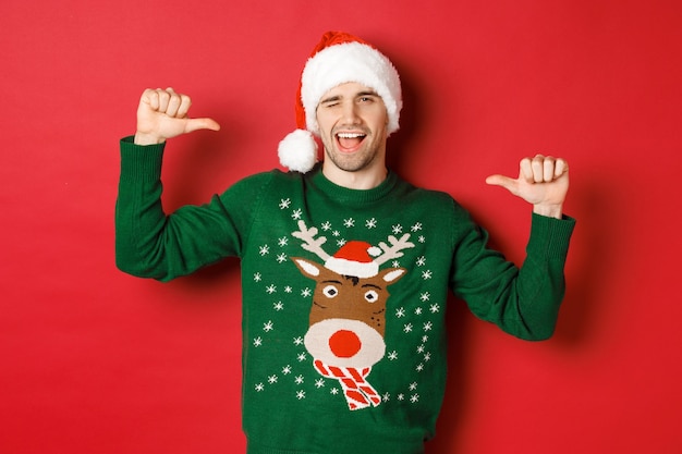 Concept of winter holidays, christmas and lifestyle. Sassy handsome man in santa hat and green sweater, pointing at himself and winking, standing over red background