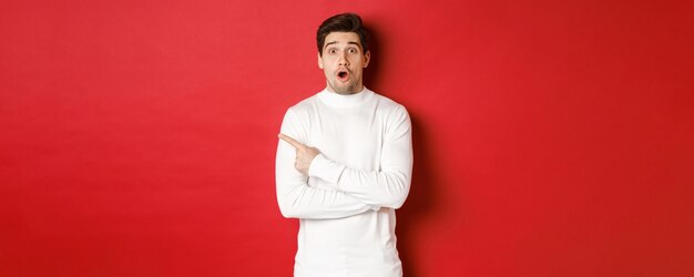 Concept of winter holidays, christmas and lifestyle. Surprised handsome guy pointing finger at new year advertisement, pointing finger left and drop jaw amazed, standing over red background.