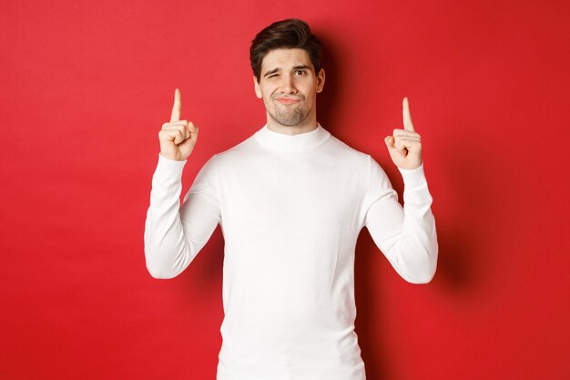 Концепция зимних праздников. Разочарованный красавчик не рекомендует промо, указывая пальцами вверх и недовольно морщась, стоя на красном фоне в белом свитере.